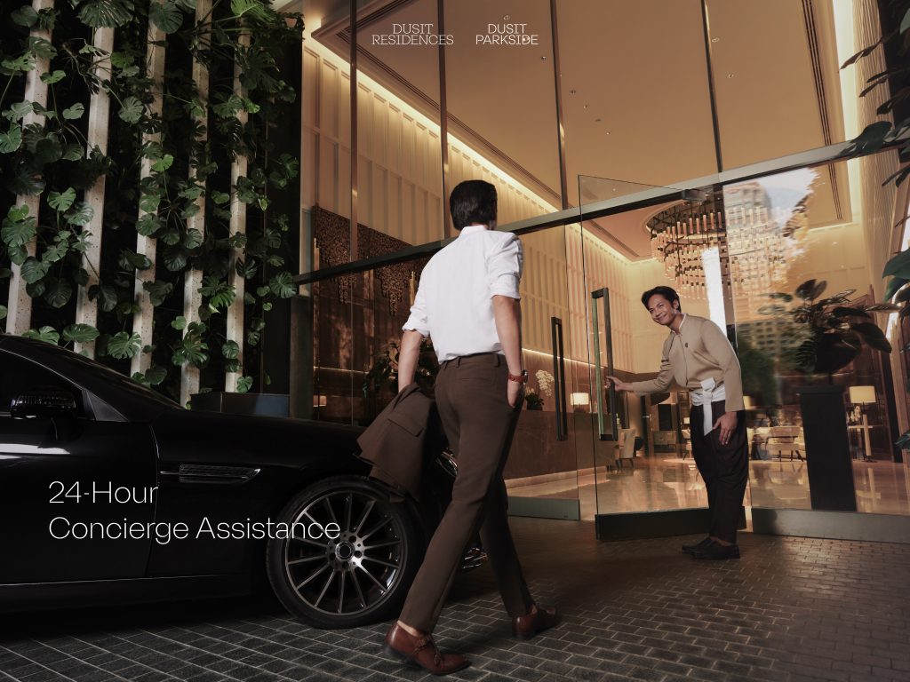 A man in business attire arrives at a luxury residence, greeted by a smiling concierge holding the glass door open. A sleek black car is parked beside a vertical garden wall. Text reads "24-Hour Concierge Assistance." ชายสวมเสื้อเชิ้ตและกางเกงสแล็คเดินเข้าสู่อาคารที่พักหรู โดยมีพนักงานคอนเซียจยิ้มต้อนรับและเปิดประตูกระจกให้ ด้านข้างมีรถยนต์สีดำจอดอยู่ริมกำแพงสวนแนวตั้ง พร้อมข้อความว่า "บริการคอนเซียจตลอด 24 ชั่วโมง"