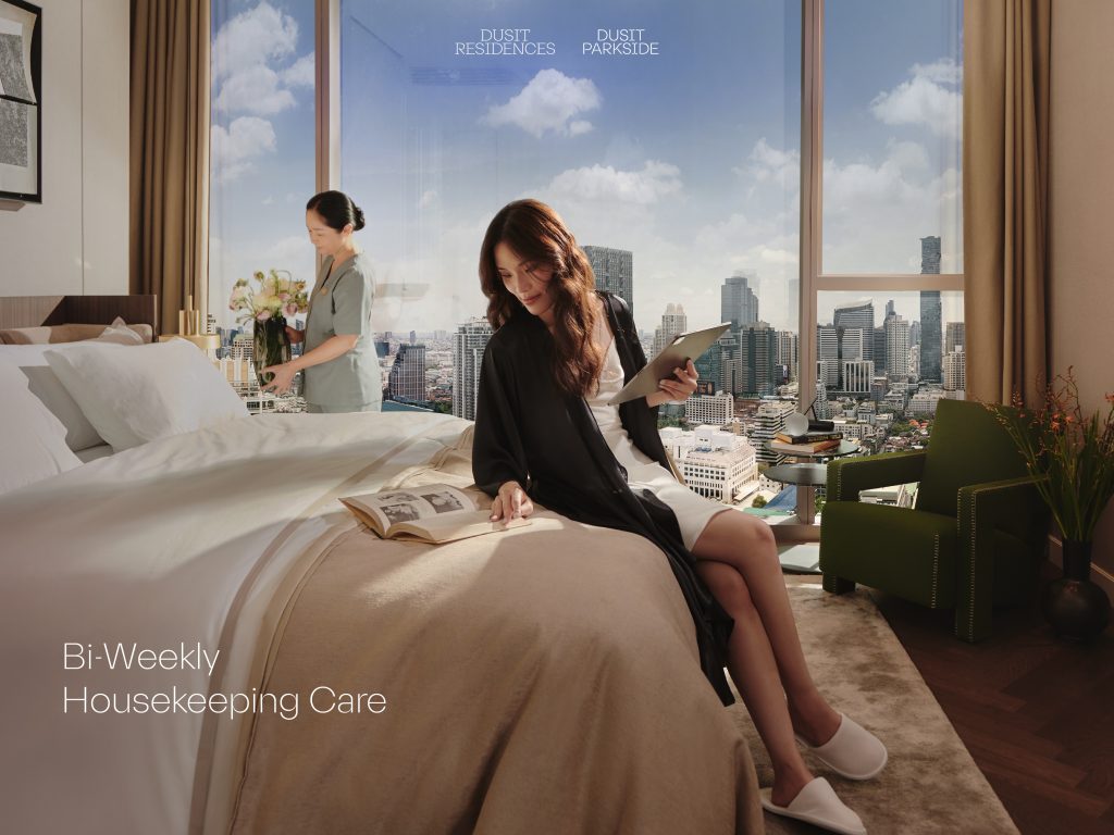 A woman in a robe relaxes on a bed with a book and tablet in hand, enjoying a panoramic city view from a high-rise bedroom. A housekeeper arranges fresh flowers nearby. Text reads "Bi-Weekly Housekeeping Care." หญิงสาวในชุดคลุมกำลังพักผ่อนบนเตียง พร้อมหนังสือและแท็บเล็ตในมือ โดยมีทัศนียภาพเมืองจากมุมสูงเป็นฉากหลัง ขณะเดียวกันพนักงานแม่บ้านกำลังจัดแจกันดอกไม้อย่างประณีต มีข้อความว่า "บริการทำความสะอาดทุกสองสัปดาห์"